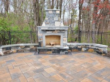 Outdoor stone fireplace with curved seating wall on a paver patio in a wooded backyard
