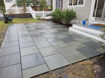 Newly installed gray paver patio beside a house with white trim and landscaping